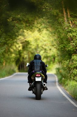 Asfalt yolda yeşil motosiklet süren siyah koruyucu takım elbiseli ve kasklı motosikletçi yeşil ağaçların arka planında, dikiz manzaralı.