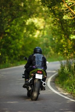 Asfalt yolda yeşil motosiklet süren siyah koruyucu takım elbiseli ve kasklı motosikletçi yeşil ağaçların arka planında, dikiz manzaralı.