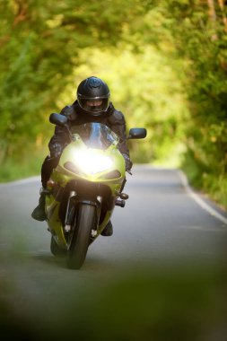 Siyah koruyucu takım elbiseli motosikletli ve kasklı yeşil motosikletli, asfalt yolda, farlar açık, arka planda yeşil ağaçlar, ön manzara