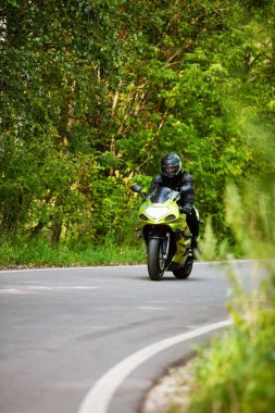 motorcyclist in black protective suit and helmet riding green motorcycle on asphalt road, headlight on, green trees in background, front view clipart