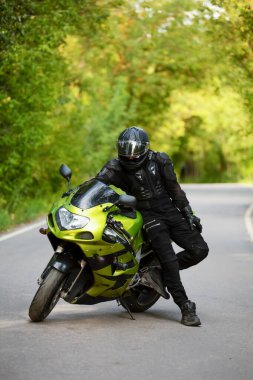 Yeşil spor motosikletinin yanında siyah koruyucu takım elbiseli ve kasklı motosikletçi, yeşil ağaçların arka planında, yan görüş,