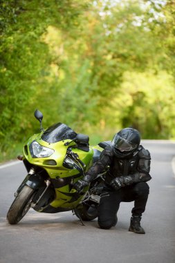 Yeşil spor motosikletinin yanında siyah koruyucu takım elbiseli ve kasklı motosikletçi, yeşil ağaçların arka planında, yan görüş,