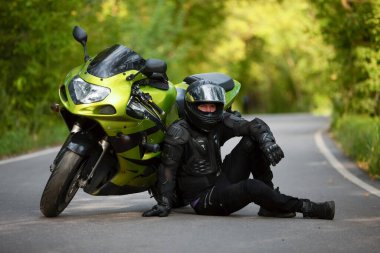 Yeşil spor motosikletinin yanında siyah koruyucu takım elbiseli ve kasklı motosikletçi, yeşil ağaçların arka planında, yan görüş,