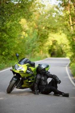 Yeşil spor motosikletinin yanında siyah koruyucu takım elbiseli ve kasklı motosikletçi, yeşil ağaçların arka planında, yan görüş,