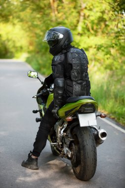 Siyah koruyucu takım elbiseli motosikletçi ve yeşil spor motosikletli kask, arka planda yeşil ağaçlar, arka manzara, yeşil bisiklet.