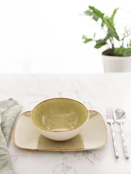用桌布 勺子和叉子放在窗前白色大理石桌子上的矩形盘子里的空绿汤碗 图库照片