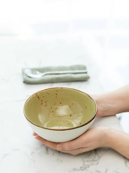 Hands holding empty green soup bowl on white marble table in front of window. Woman hands holding empty soup bowl