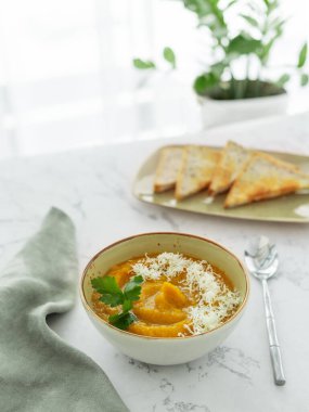 Bir kase balkabağı ya da taze maydanozla süslenmiş havuçlu vejetaryen kremalı çorba ve rendelenmiş çedar peynirli beyaz mermer masa. Taze ekmek tostları ve masa örtüsüyle.