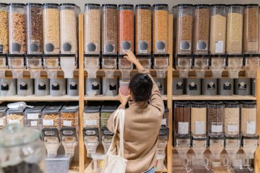 Afro-Amerikalı kadın, sıfır atık marketinde tekrar kullanılabilir paketleri toplu ürünlerle dolduruyor. Sürdürülebilir eko marketten tahıl ve mısır gevreği alan kadının arka görünüşü.