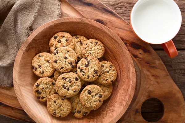 Chocolate chip cookies in wooden bowl with a cup of milk on the side, photographed overhead on wood (Selective Focus, Focus on the cookies on the top)