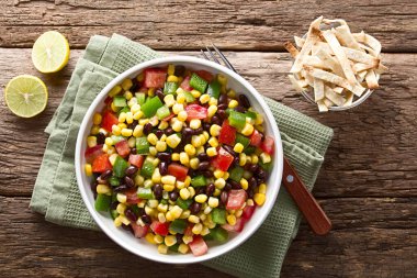 Mexican style colorful fresh vegetable salad made of beans, corn, tomato and bell pepper served in bowl, baked tortilla strips, limes and fork on the side, photographed overhead on wood (Selective Focus, Focus on the salad)