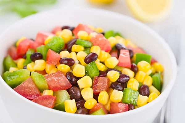 Mexican style colorful fresh vegetable salad made of beans, corn, tomato and green bell pepper served in white bowl, photographed on white wood (Selective Focus, Focus one third into the salad)