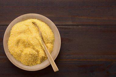 Raw coarsely ground cornmeal or polenta on wooden plate with wooden spoon, photographed overhead on dark wood with copy space on the side (Selective Focus, Focus on the cornmeal)