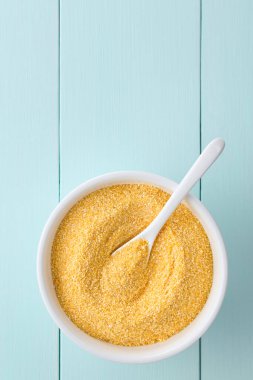 Raw coarsely ground cornmeal or polenta in white bowl with spoon, photographed overhead on blue wood with copy space above (Selective Focus, Focus on the cornmeal)