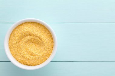 Raw coarsely ground cornmeal or polenta in white bowl, photographed overhead on blue wood with copy space on the side (Selective Focus, Focus on the cornmeal)