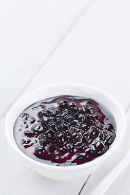 Fresh homemade jam made of Patagonian Calafate berries (lat. Berberis heterophylla), served in white bowl, photographed on white wood with copy space above (Selective Focus, Focus one third into the jam)