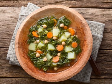Fresh homemade vegan vegetable soup made of kale, chickpea, carrot, celery, onion and potato served in wooden bowl, photographed overhead on wood (Selective Focus, Focus on the soup)