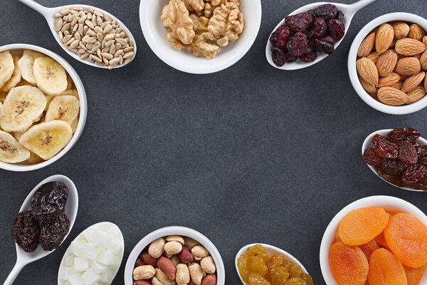 Variety of dried fruits, nuts and seeds (sunflower seed, walnut, cranberry, almond, raisin, apricot, sultana, peanut, coconut, prune, banana) in small bowls, photographed overhead on slate with copy space in the middle