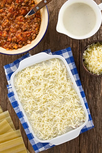 Grated cheese on top of the unbaked homemade lasagna, ingredients on the side, photographed overhead on wooden table (Selective Focus, Focus on the grated cheese in the casserole dish)