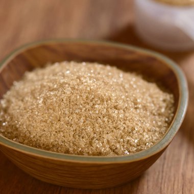 Kahverengi şeker kristalleri küçük kasede, ahşap tahta üzerinde doğal ışık ile fotoğraflanmış ve arkada ölçüm kaşığı (Çok Sığ Saha Derinliği, kasenin üçte biri kadar odaklanın)