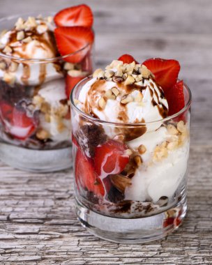 Summer cold dessert - Plombir ice cream scoop with chocolate sauce, cookie crumbs, ripe strawberries, crushed peanut nuts in a glass on a wooden table