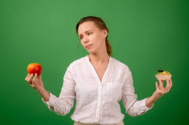 Elmalı ve pastalı orta yaşlı bir kadın kendi seçimiyle eziyet görüyor. Yeşil bir arka planda. Doğru sağlıklı yiyeceği seçme fikri..
