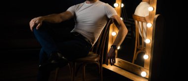 Beyaz tişörtlü, kolunda saat olan tanınmamış bir adam aynanın yanındaki sandalyede oturuyor..