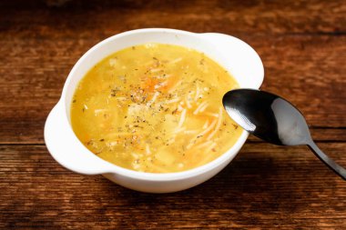 Erişteli tavuk çorbası, patates, soğan ve havuç, ahşap arka planda beyaz bir tabakta..