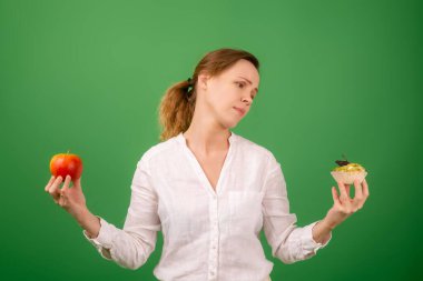 Elmalı ve pastalı orta yaşlı bir kadın kendi seçimiyle eziyet görüyor. Yeşil bir arka planda. Doğru sağlıklı yiyeceği seçme fikri..