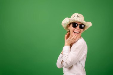 Yeşil arka planda beyaz gömlekli, şapkalı ve güneş gözlüklü neşeli bir kadın. Metnin için yer.