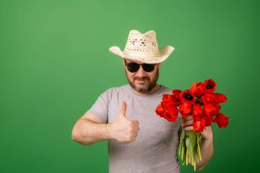 A man in a hat and sunglasses shows an approving gesture and holds a bouquet of tulip flowers on a green background. Romanticism, give, apologize concept.