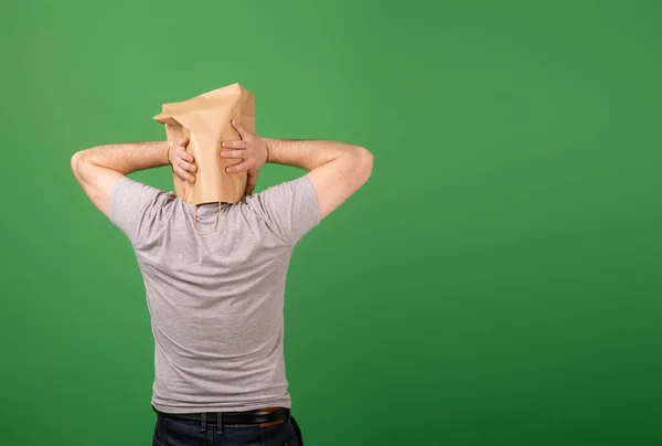 An unrecognizable man with a paper bag on his head grabbed his head against a green background. Place for your text. mental health, greenwashing, disinformation concept