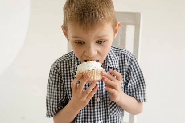 A young boy in a shirt eats a cake on a light background. The food joy of sweets.