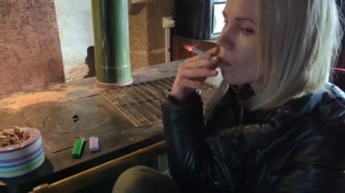 Siyah ceketli orta yaşlı bir kadın içeride sigara içiyor. Yavaş çekim.