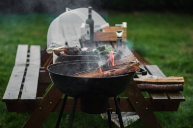The smoking brazier of a round form. Firewood flares up in the brazier against the background of a table and chairs. The concept of picnic, relaxation, grilling.