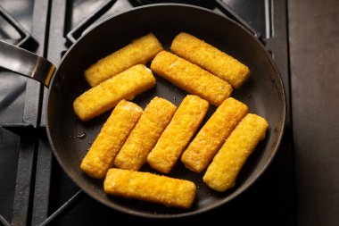 Breaded fish sticks in a frying pan. Preparation of frozen fish sticks. Fast food. Dark background.