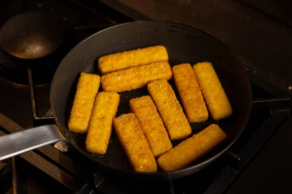 Breaded fish sticks in a frying pan. Preparation of frozen fish sticks. Fast food. Dark background.