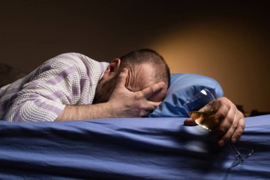 A man with a headache holds a glass of wine in bed. Alcoholism, loneliness, hangover concept. A dark room.