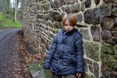 Sad boy near the stone wall. A boy of nine years old in a warm jacket stands with his head bowed. A child experiencing negative emotions.