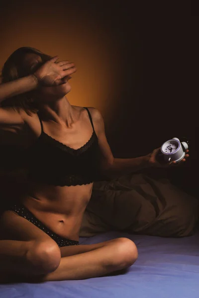 A woman on the bed in the morning holds an alarm clock in her hands in the dark. Poor sleep, violation of the regimen, lack of sleep concept. A middle aged woman in her forties is sitting on a bed trying to wake up.