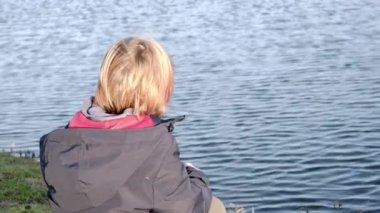 The thoughtfulness of an unrecognizable child in front of a pond. View from the back. School-age boy with long red hair.