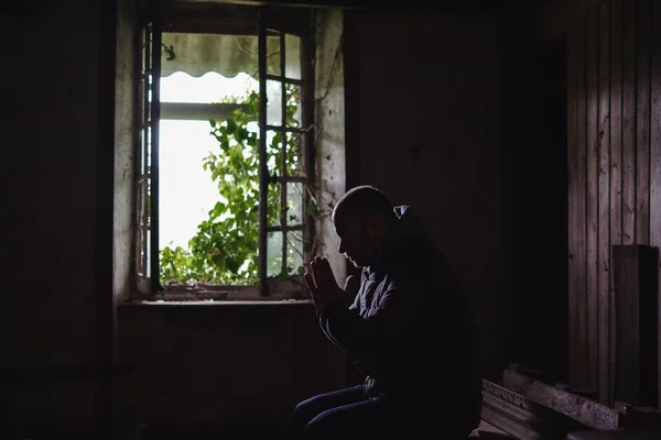 A man prays in an old crumbling house. Abandoned house with an open window from the inside. Hope, despair concept.