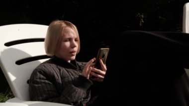 Woman communicates by video call outdoors. A middle-aged woman in her forties sits on a chair in the backyard of a house.