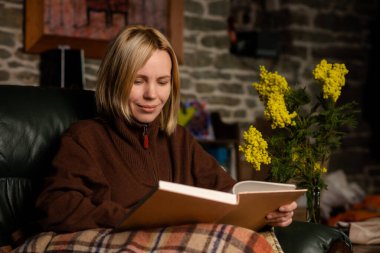 A middle-aged woman, forty years old, is reading a book on the couch. Woman in warm clothes covered with a blanket. Dark key photography.