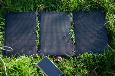 Portable solar battery and mobile phone on green grass. Charging electronic gadgets on a hike, during outdoor recreation. Ecological energy concept.
