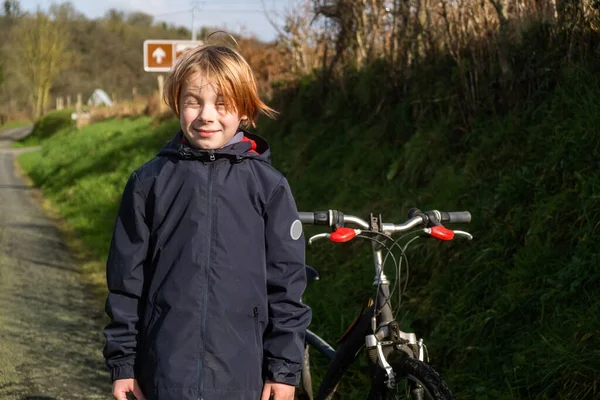 Young boy with bicycle in countryside. Boy of nine or ten years of old age stands on green grass near road. There is bike nearby. Active recreation, cycling.