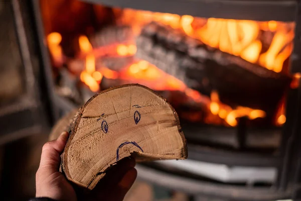 Rise in price of firewood for heating concept. Hand holds log with sad smiley against background of burning fireplace