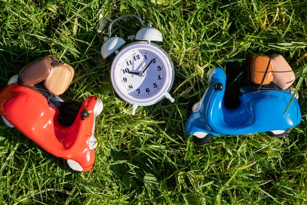 Toy scooters and alarm clock on green grass. Delivery, travel concept.