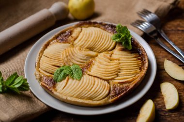 Ağız sulandıran, ahşap yüzeye yerleştirilmiş Fransız turtasının görüntüsü. Pasta taze nane yapraklarıyla süslenir ve üzerine mis kokulu tazelik katılır. İnce dilimlerle kaplı kıtır kıtır elmalar üzerine baharat ve şeker serpiştirilmiş..