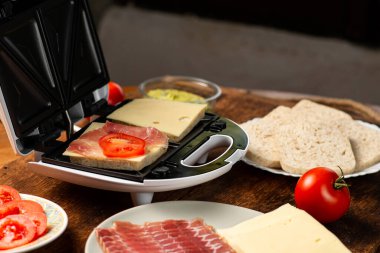 Sandviç makinesinde sandviç pişirmek. Farklı malzemeler. Ekmek, peynir, ahşap arka planda sebze..